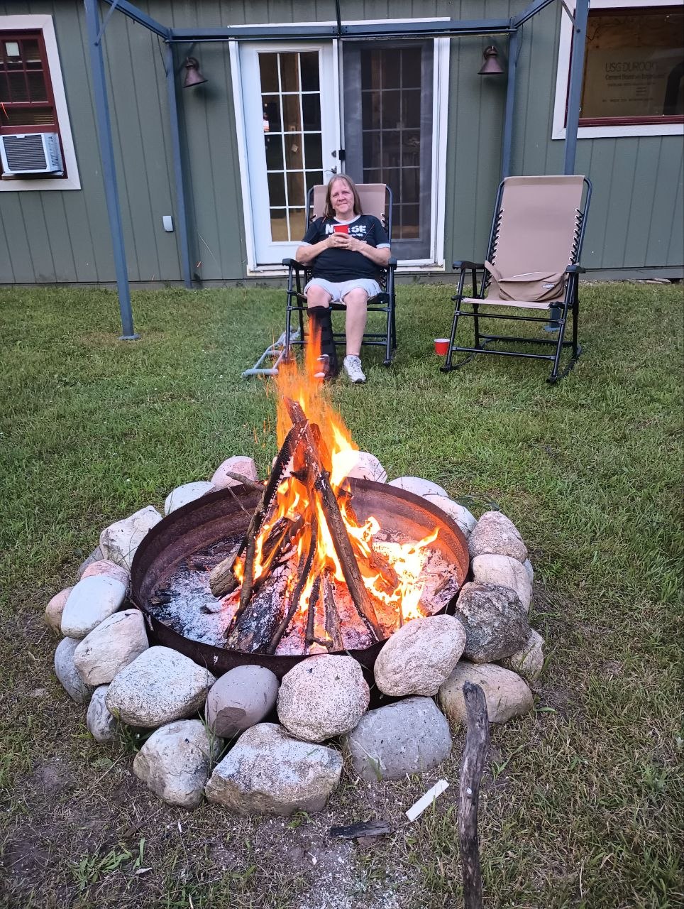 Vanessa relaxing by a later-season bonfire in front of the more settled cabin, with her walking cast faintly visible